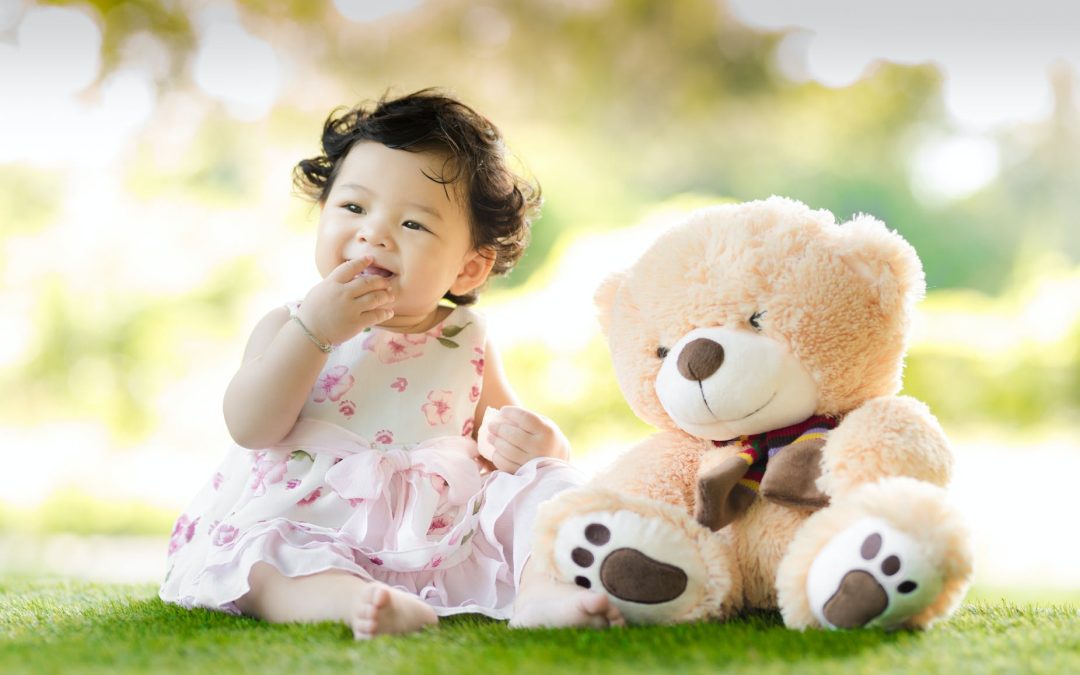baby sitting on green grass beside bear plush toy at daytime